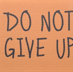 Cardboard sign reading 'Do Not Give Up' placed on green turf, symbolizing perseverance.